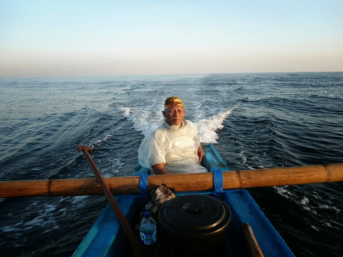 A Balinese fisherman steers a wooden outrigger boat across the open sea in morning light