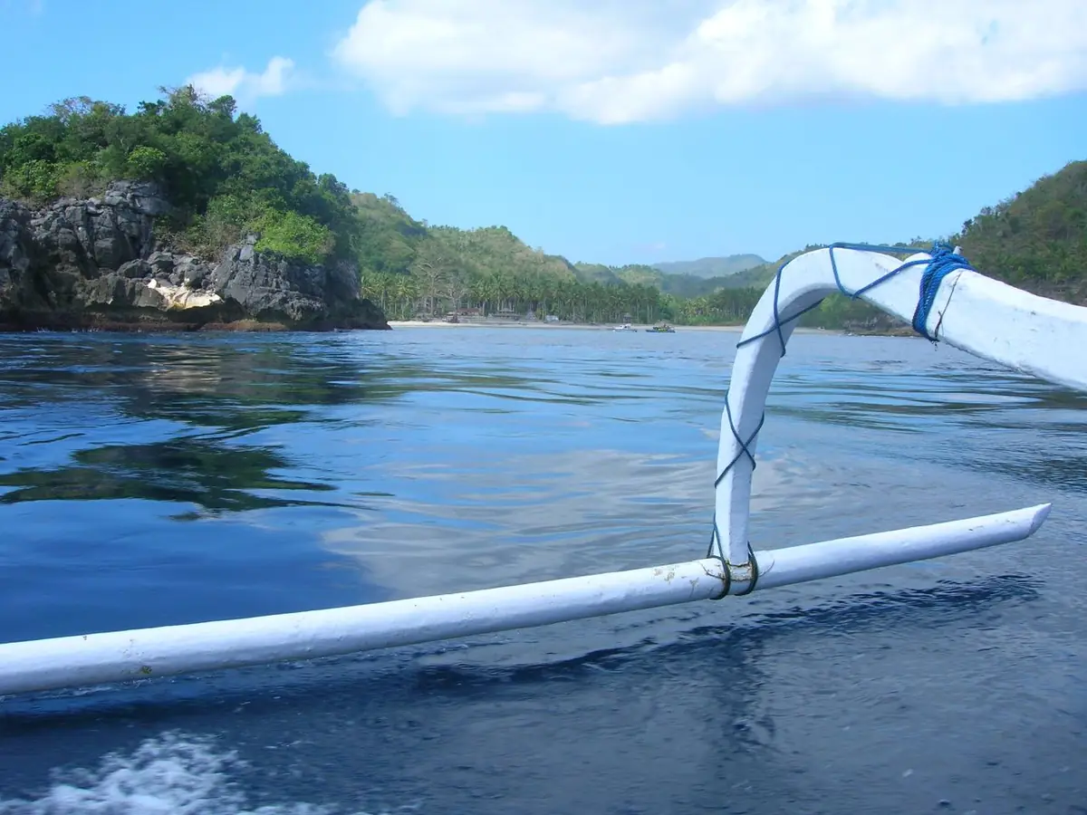 The bamboo outrigger of a traditional boat on deep blue water below green cliffs