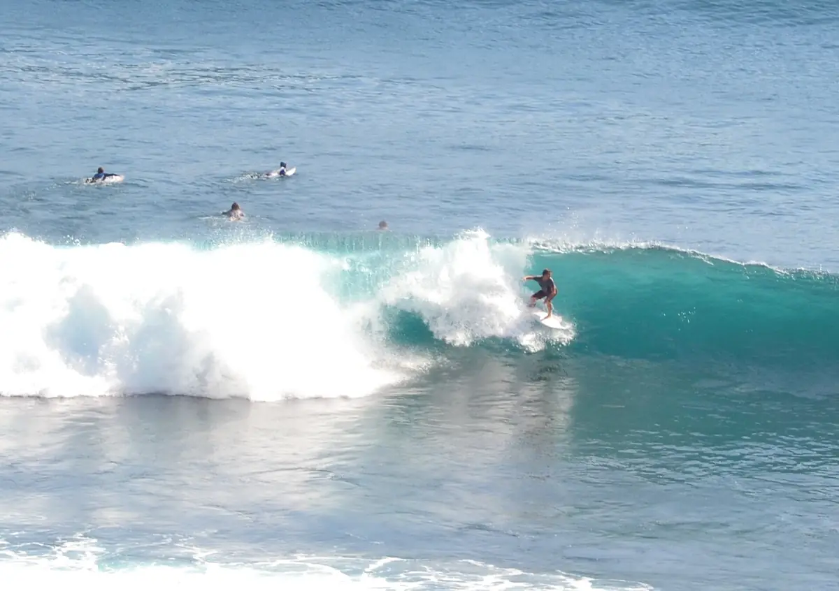 A surfer rides along a turquoise wave in Bali while others wait in the line-up